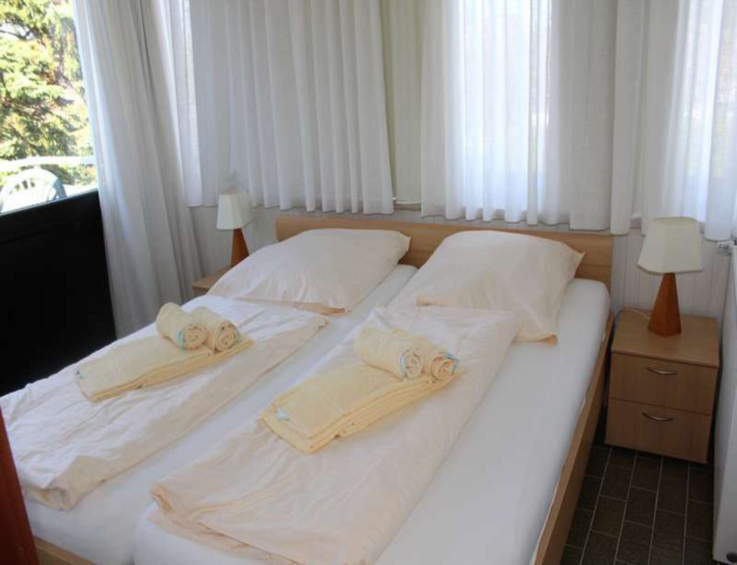 A double bed with white linens, two bedside tables with lamps, and a window with white curtains.