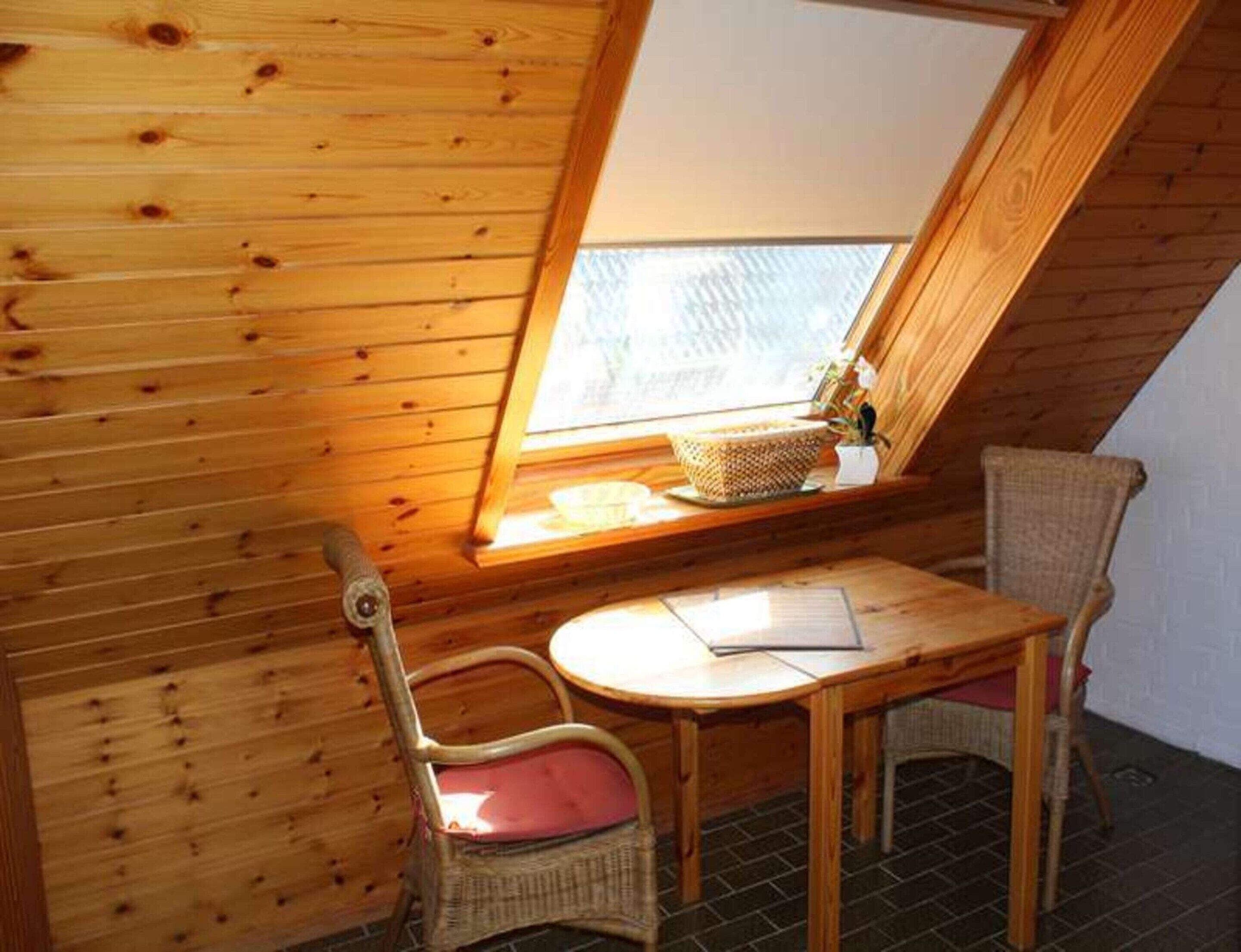 Et lille rum med skråvæg, trævægge, et vindue, et bord, to stole og en kurv på vindueskarmen.