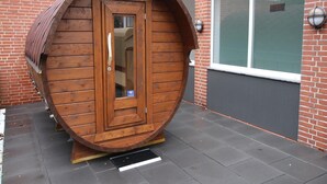 A wooden barrel structure with a door, positioned on a paved surface with a window and a brick wall in the background.