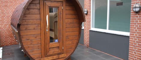 A wooden barrel structure with a door, positioned on a paved surface with a window and a brick wall in the background.