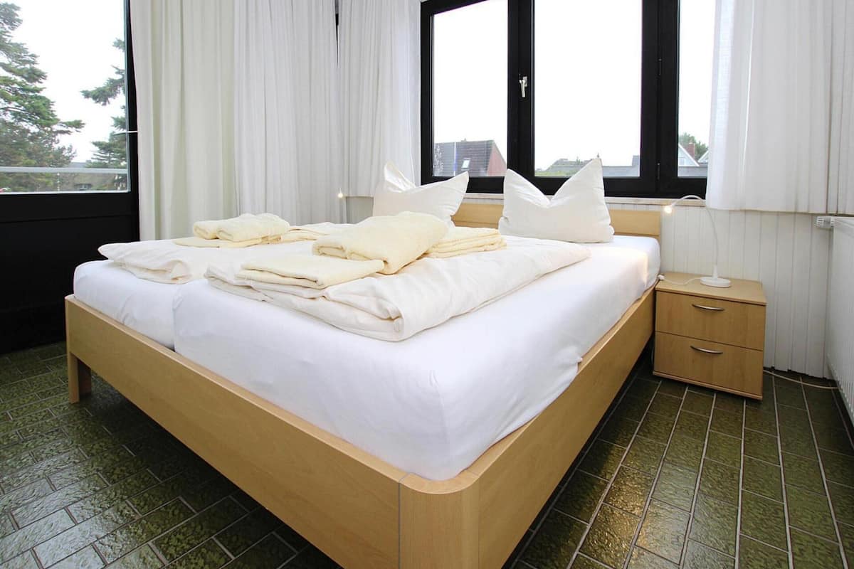 A neatly made bed with white linens and pillows, a bedside table with a lamp, and a window with curtains.