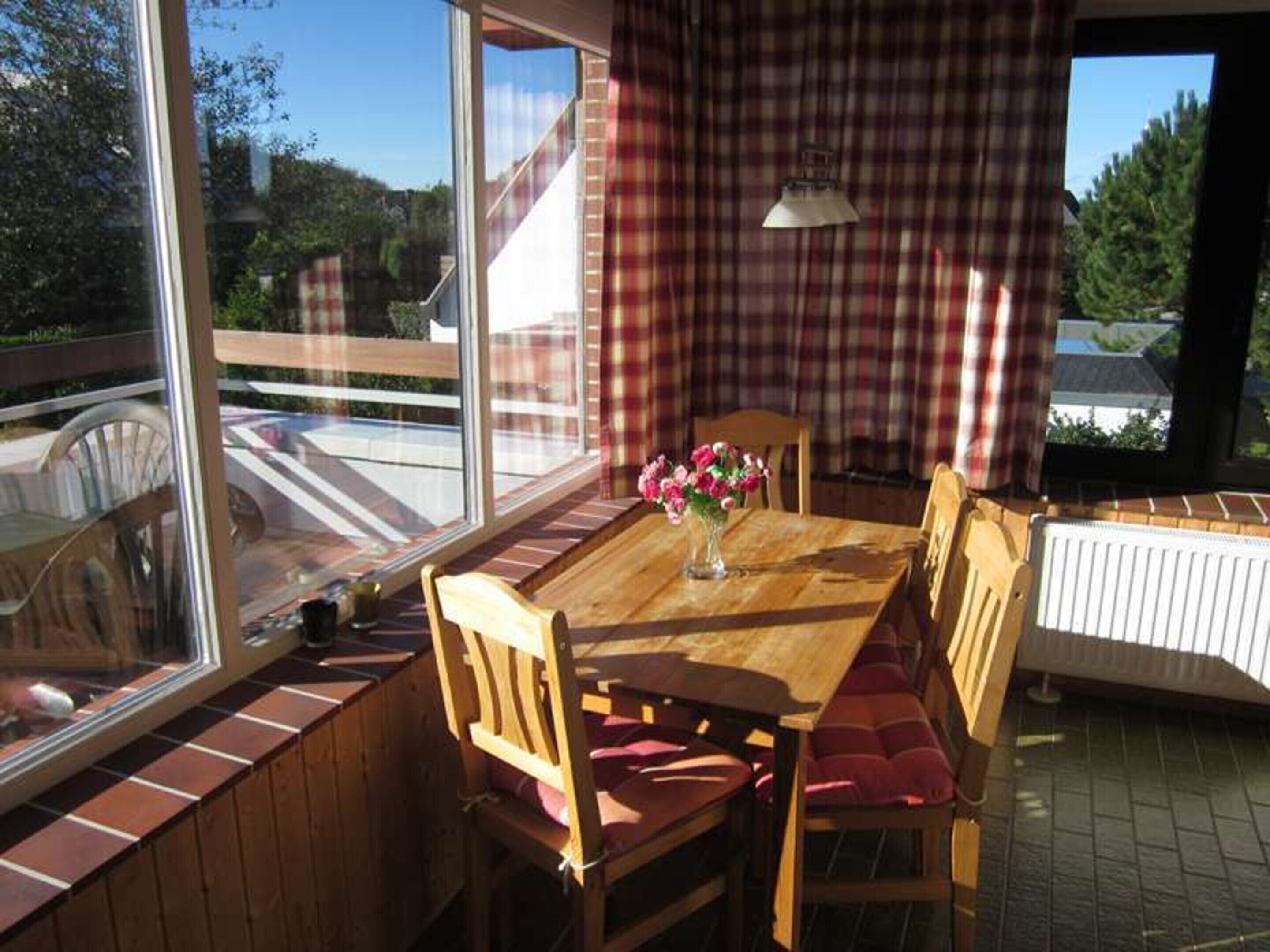 Une table de salle à manger en bois, entourée de chaises rembourrées de couleur rouge, un vase rempli de fleurs et une fenêtre donnant sur un quartier résidentiel.