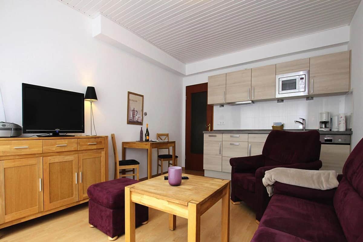 A modern living room with a wooden TV stand, a purple sofa, a wooden coffee table, and a kitchen area with a microwave and cabinets.