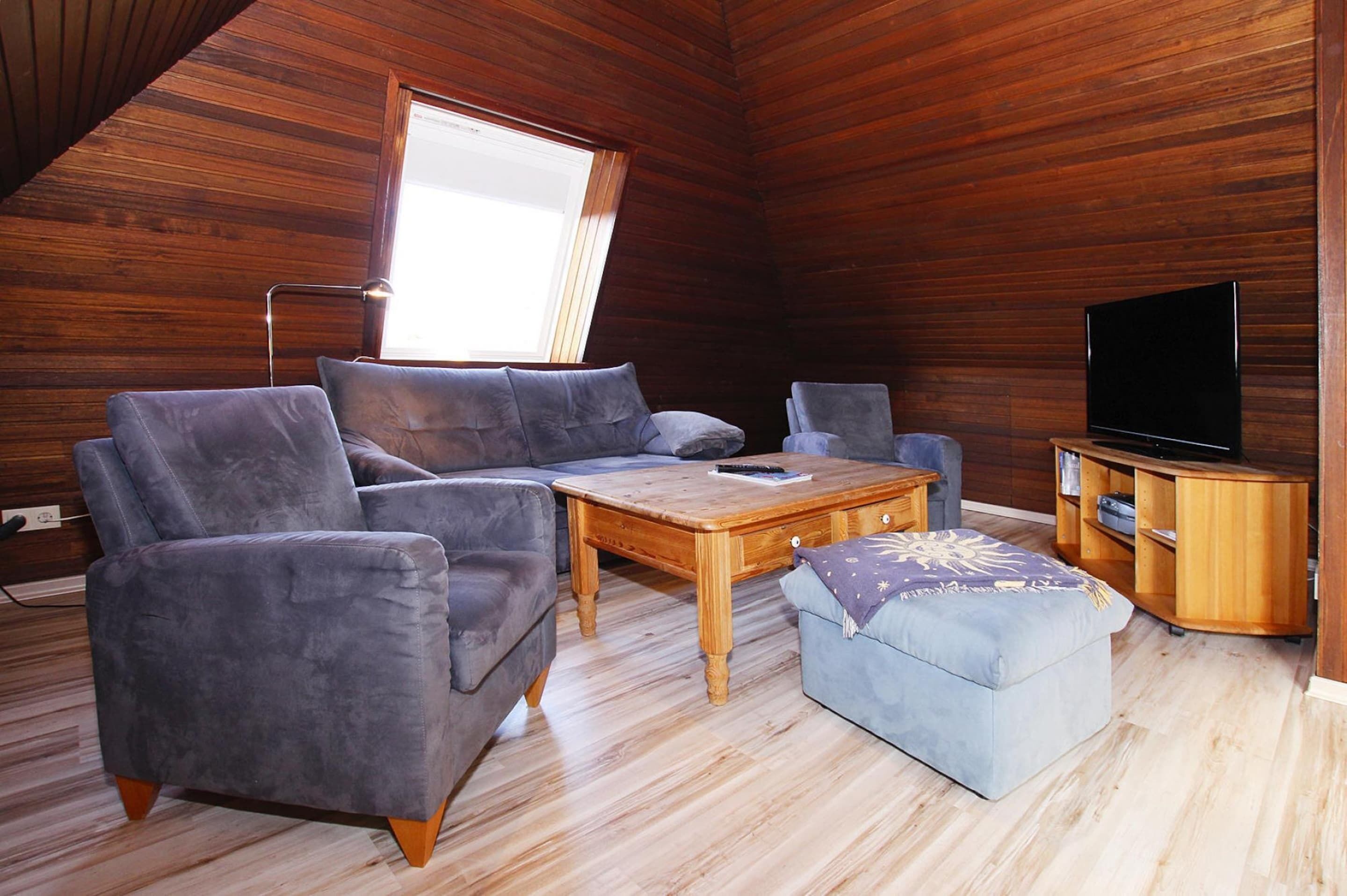 A cozy room with a wooden ceiling, a sofa, a coffee table, a TV stand, and a window.