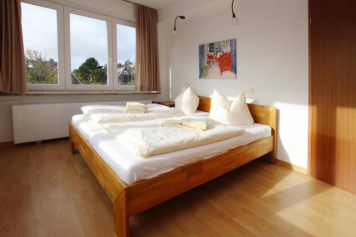A bedroom with a wooden bed, white bedding, a radiator, a window with curtains, and a painting on the wall.