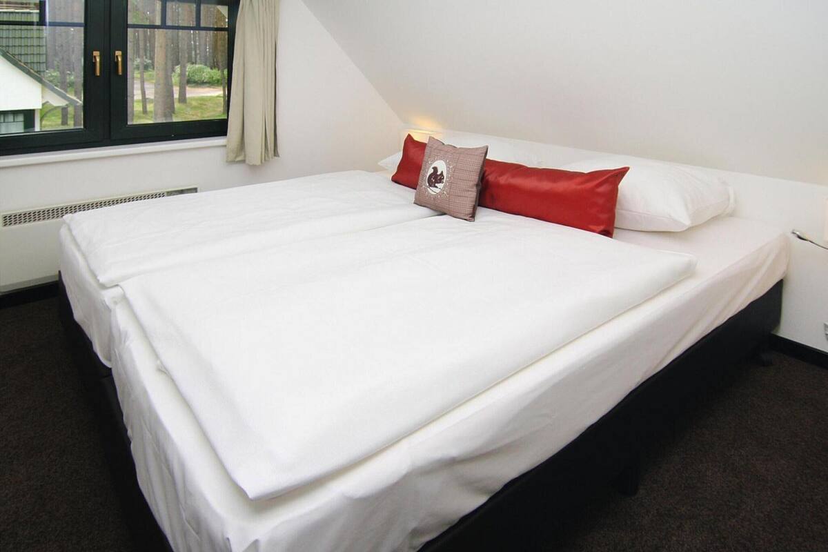 Un lit bien fait, avec du linge de lit blanc et deux oreillers, un rouge et l’autre orné d’un logo, dans une pièce dotée d’une fenêtre et d’un radiateur.