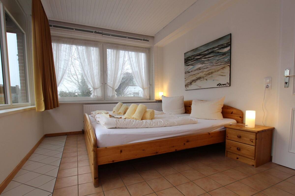 A bedroom with a wooden bed, white bedding, a nightstand, a lamp, and a painting on the wall.