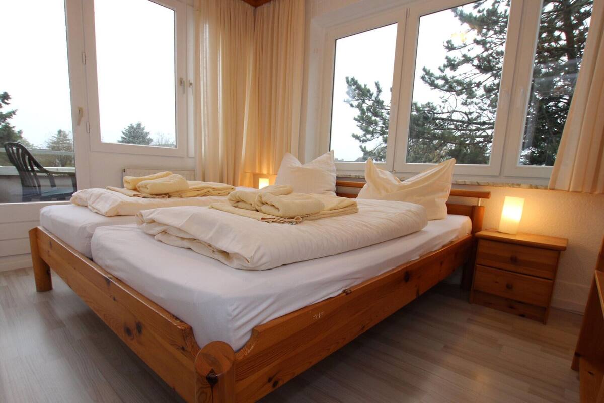 A wooden bed with white linens and pillows, a bedside table with a lamp, and large windows with curtains.