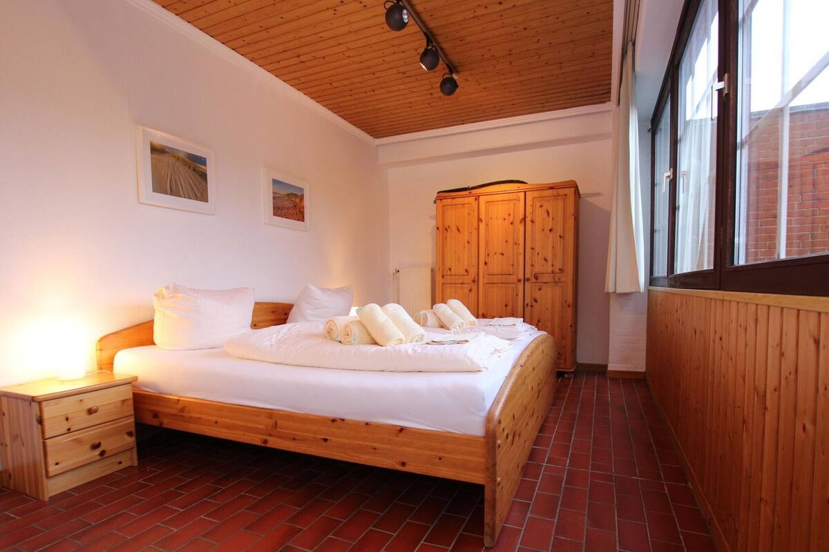 A wooden bed in a room with a brick floor, a wooden wardrobe, and a window with curtains.