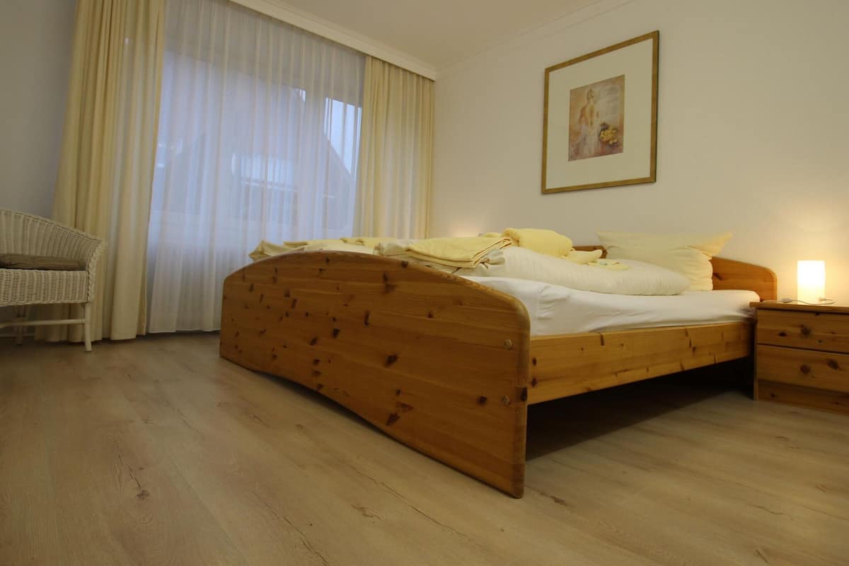 A bedroom with a wooden bed, a nightstand, a wicker chair, and a large window with sheer curtains.