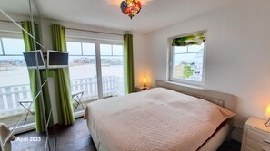 Habitación de hotel con cama, escritorio, silla, televisor y balcón con vista a la playa y edificios.
