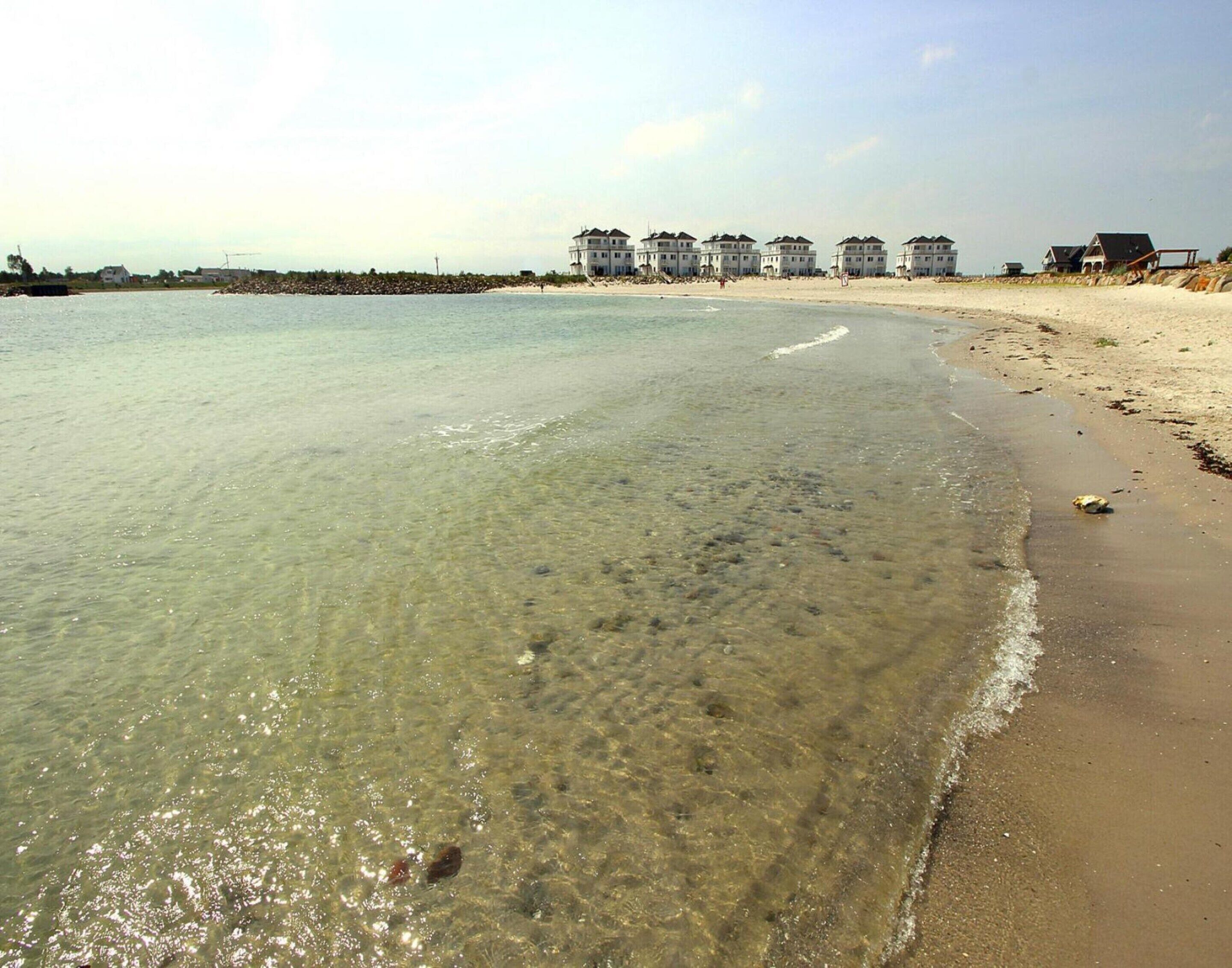 A beach with clear shallow waters, a sandy shore, and a row of white buildings in the background.