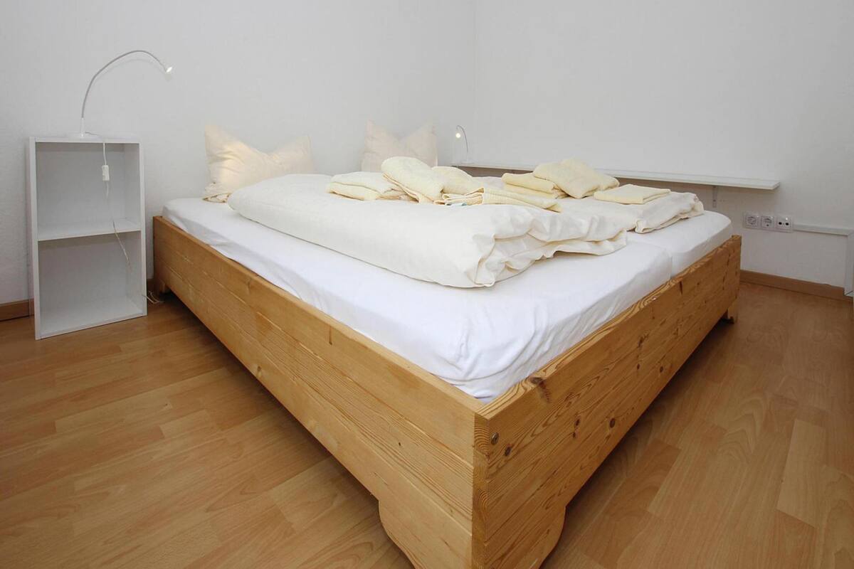 A wooden bed with white bedding in a room with white walls and a white shelf.
