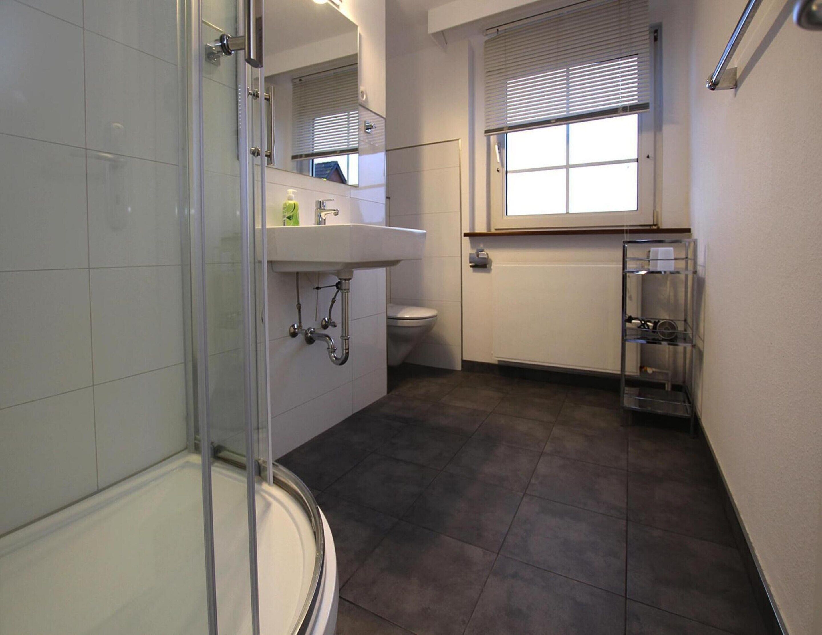 Un baño con cabina de ducha de vidrio, lavamanos blanco, inodoro y ventana con persianas.