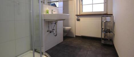 Un baño con cabina de ducha de vidrio, lavamanos blanco, inodoro y ventana con persianas.