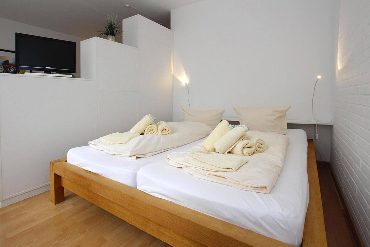 A modern bedroom with a wooden bed, white linens, and a flat-screen TV on a white wall.