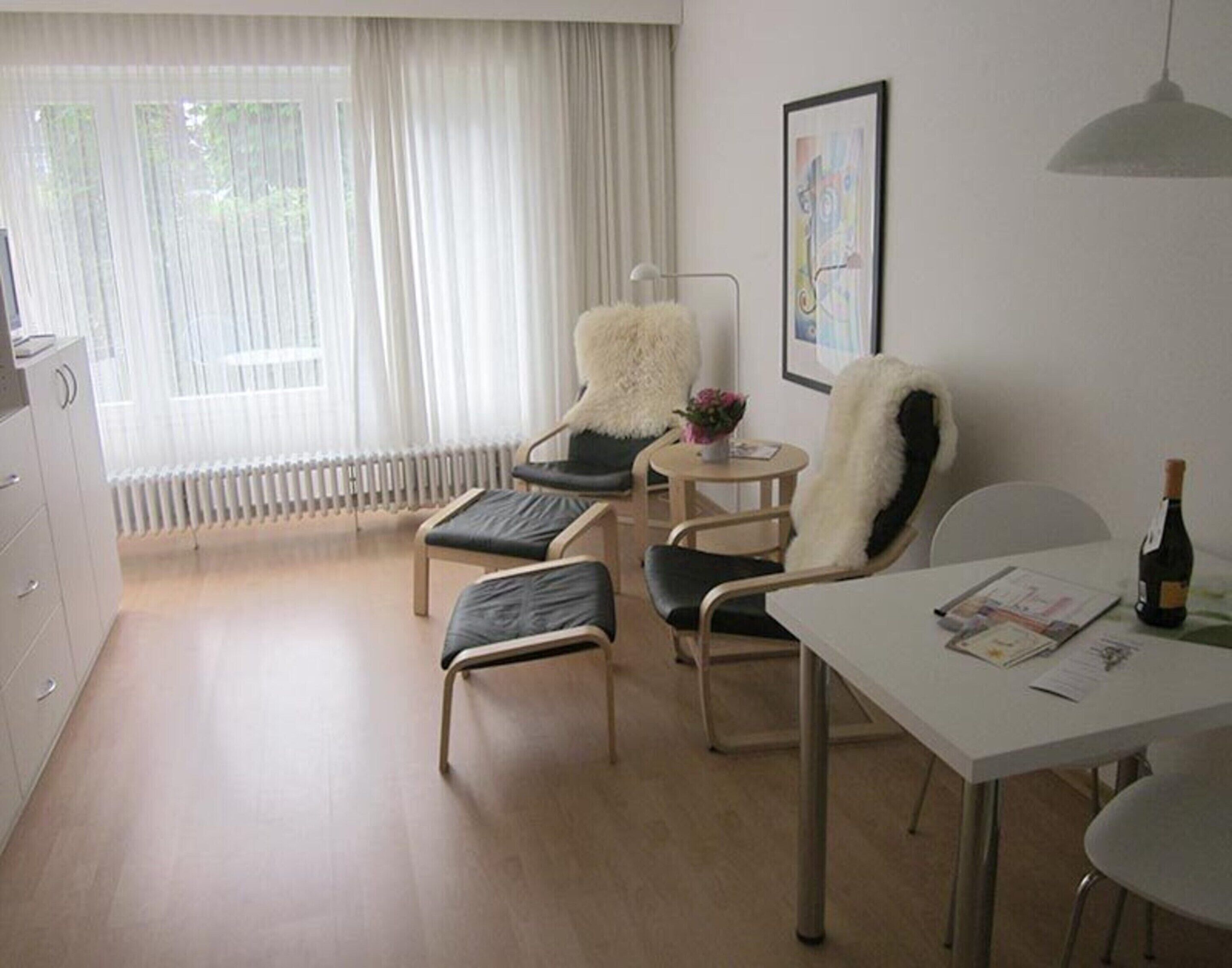 A modern, well-lit room with a dining table, two chairs, a small round table with a vase, and a radiator under the window.