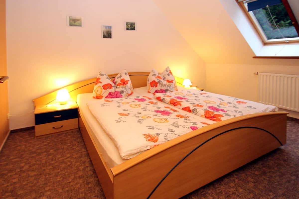 A bedroom with a wooden bed, floral bedding, a nightstand, a lamp, and a small window with a view of trees.
