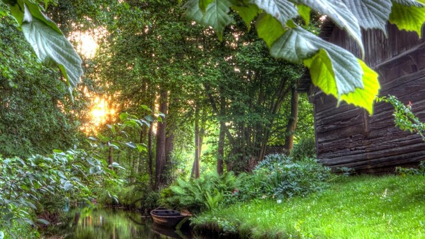 A serene riverside scene with a wooden boat, lush greenery, and a rustic building. - Apartments Spreewaldperle, Alt Zauche (Alt Zauche-Wußwerk)