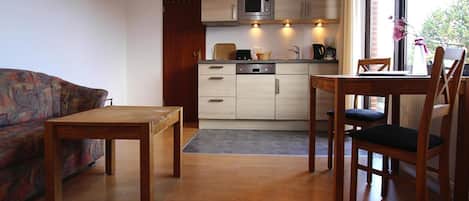 A compact living space with a sofa, a wooden table, a kitchen area with a microwave, and a window with a view of greenery.