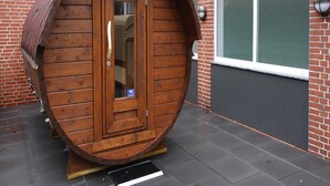 A wooden barrel-shaped structure with a door, placed on a paved surface next to a brick building with a window. - Apartment House Katharina, Westerland (Sylt)