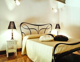 Una cama bien hecha con estructura metálica, dos lámparas de noche, una mesita auxiliar pequeña y una pared lisa.