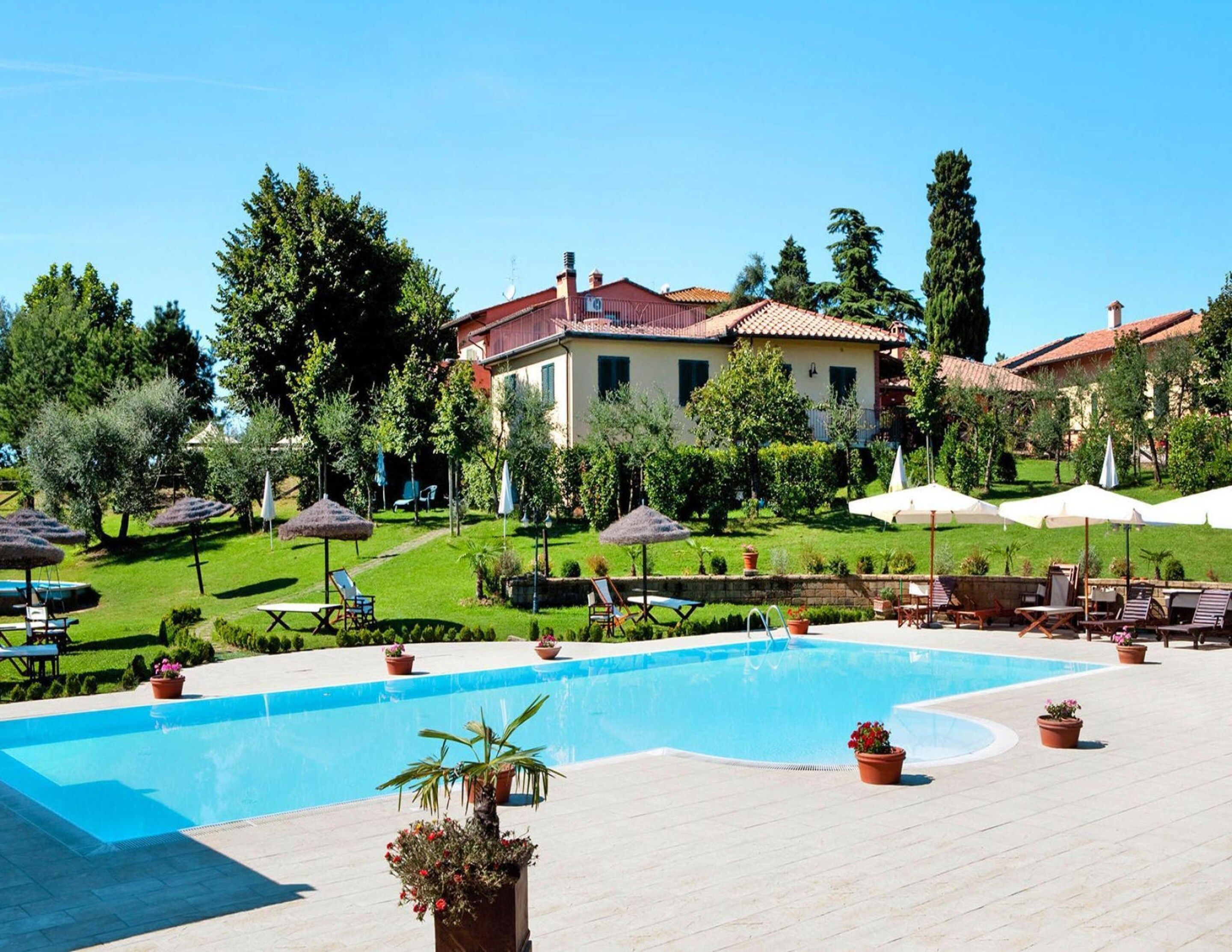 A swimming pool with lounge chairs and umbrellas, surrounded by greenery and a building with a tiled roof.