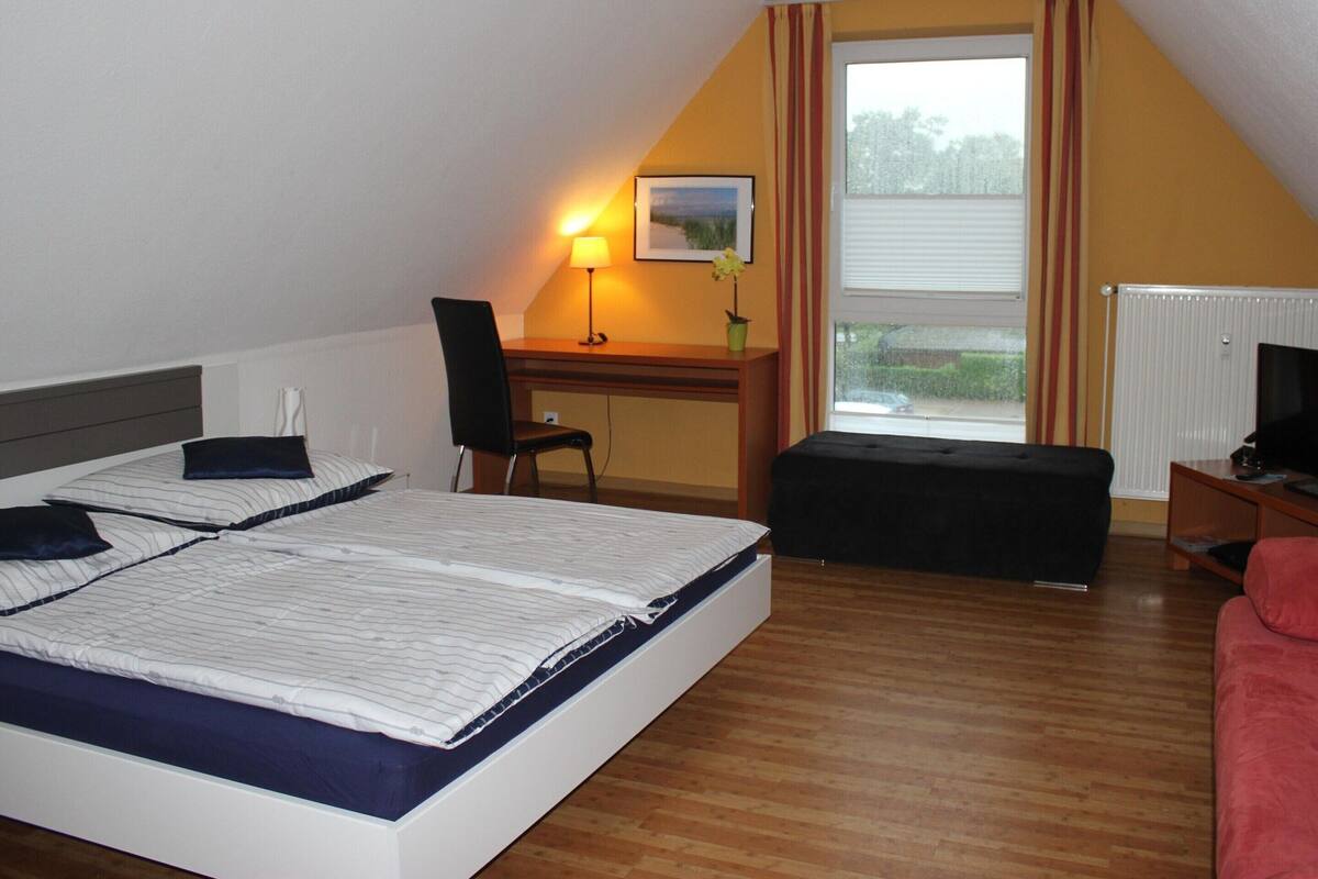 A hotel room with a large bed, a desk with a chair, a small table with a lamp, a window with curtains, and a radiator.