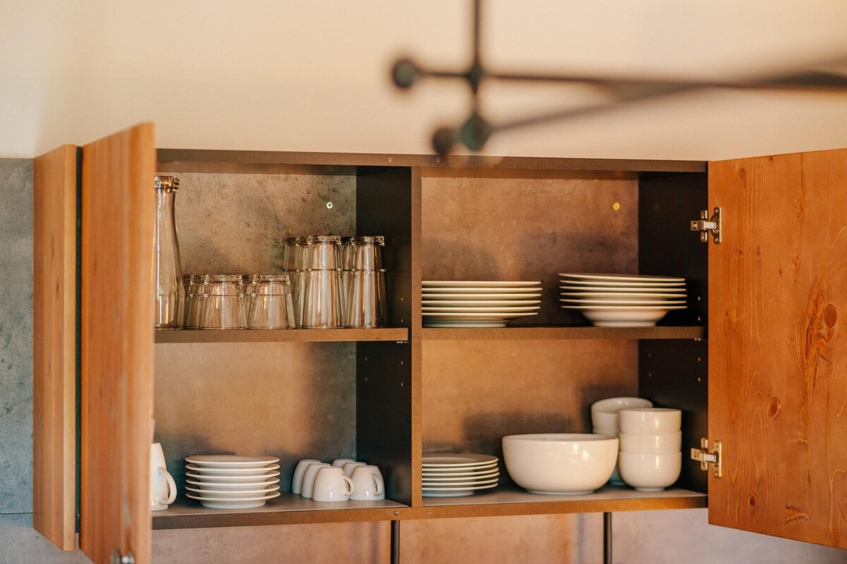 A kitchen cabinet with open shelves containing glass jars, plates, bowls, and mugs.