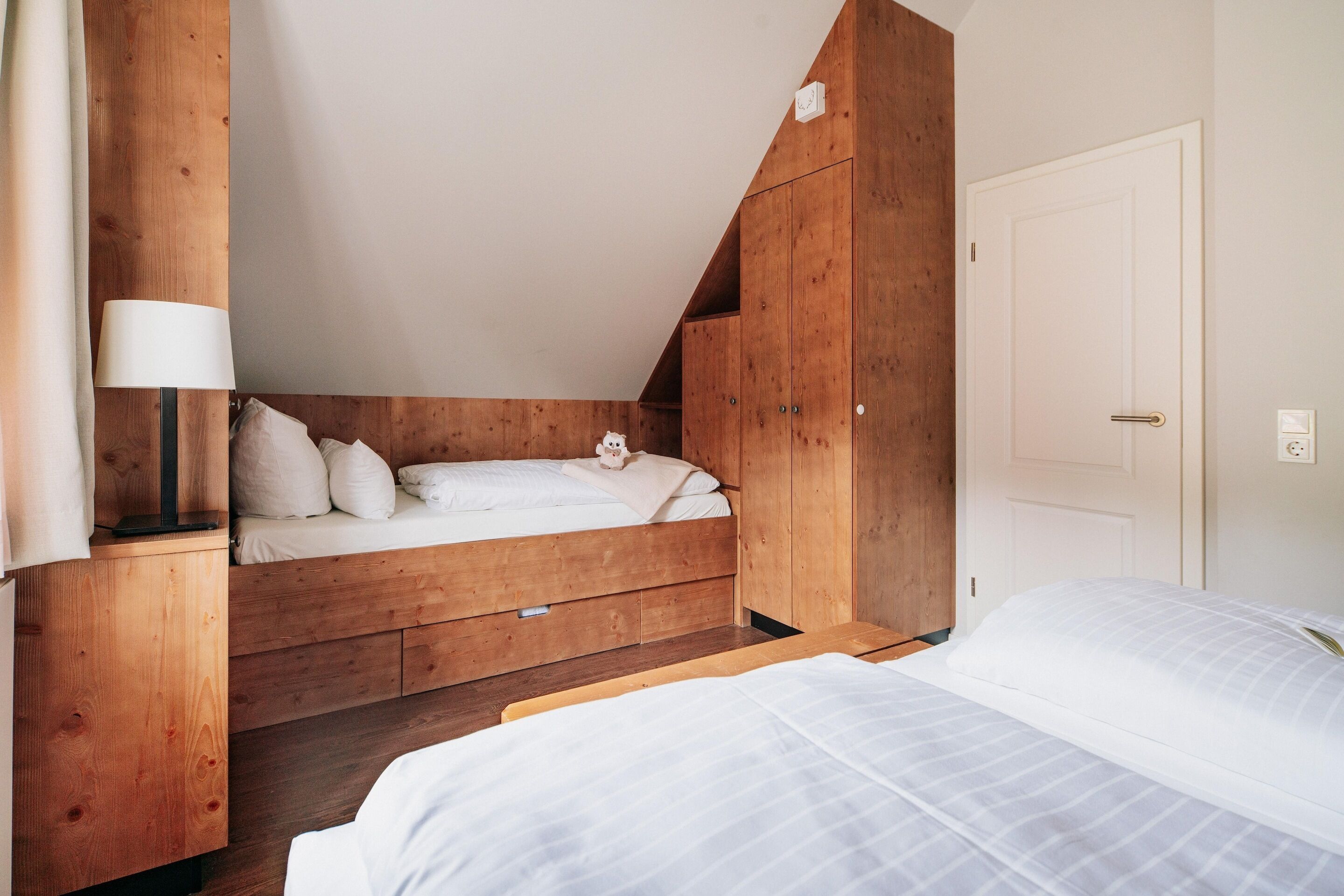 A bedroom with a wooden bunk bed, a bedside table with a lamp, and a white door.