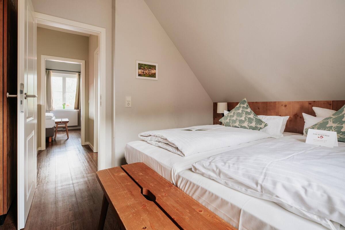 A bedroom with two beds, a wooden bench, a desk, and a window with curtains.