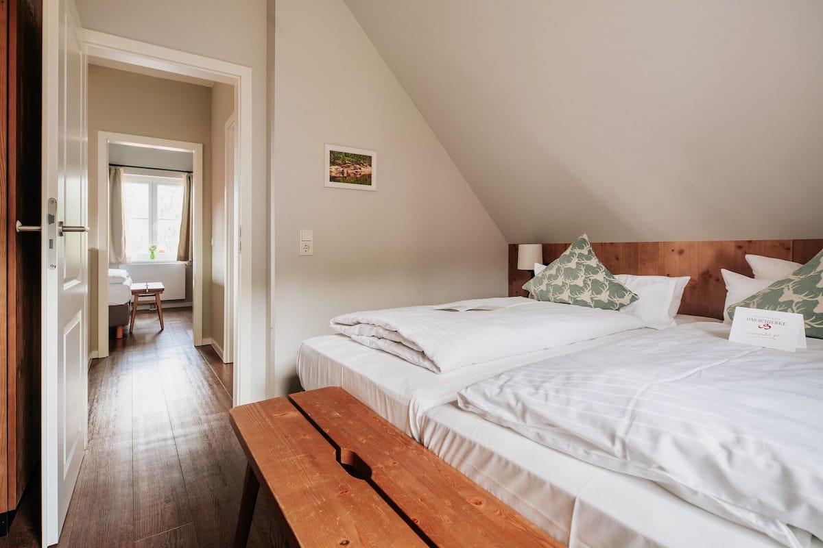 A bedroom with two beds, a wooden bench, a desk, and a window with curtains.