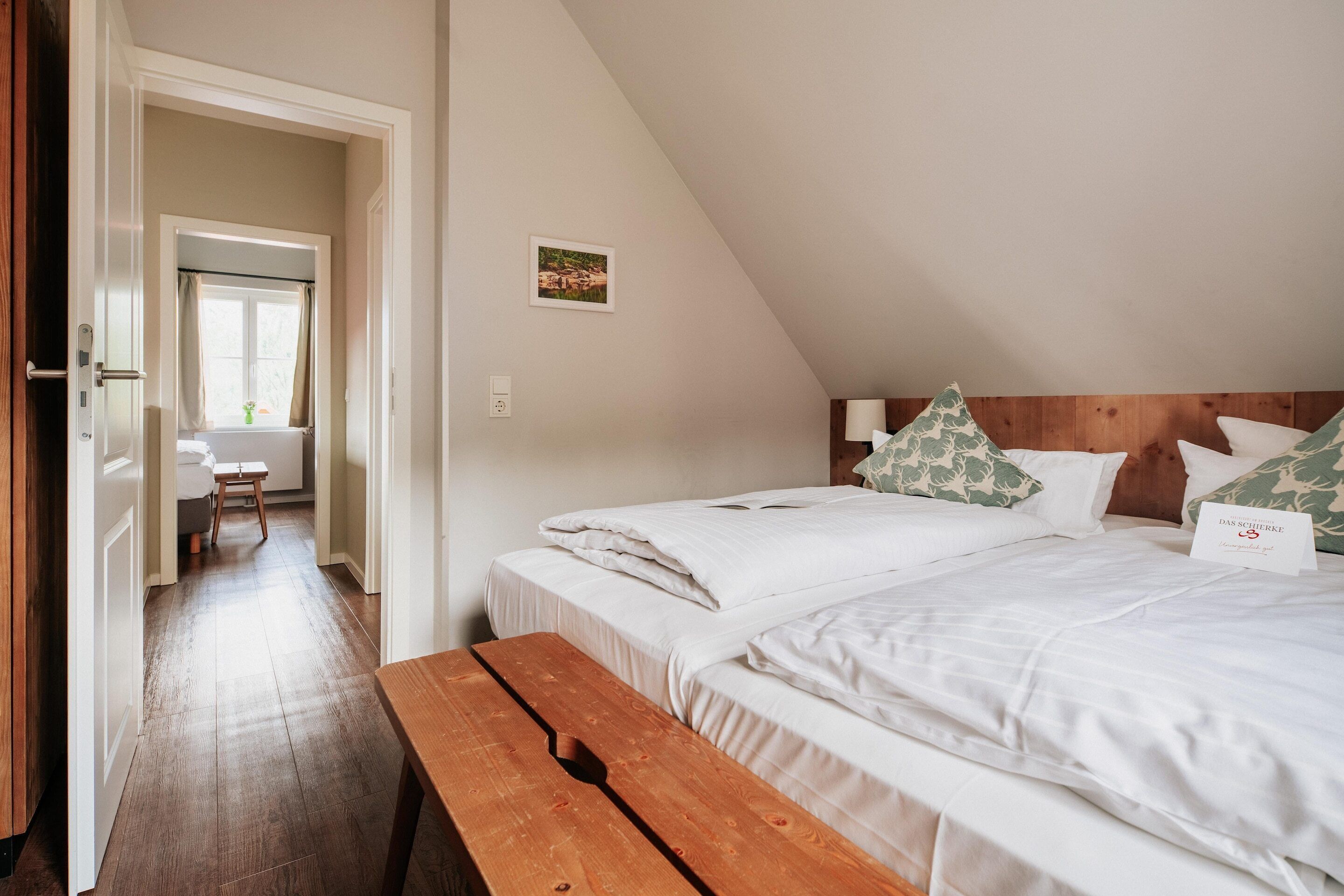A bedroom with two beds, a wooden bench, a desk, and a window with curtains.