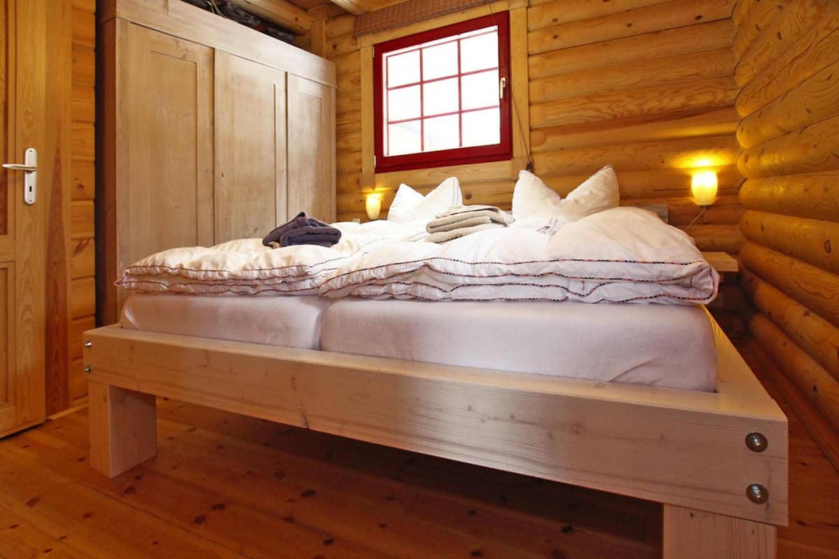 Une pièce de cabane en bois comprenant un lit, une fenêtre à cadre rouge et des lampes fixées au mur.