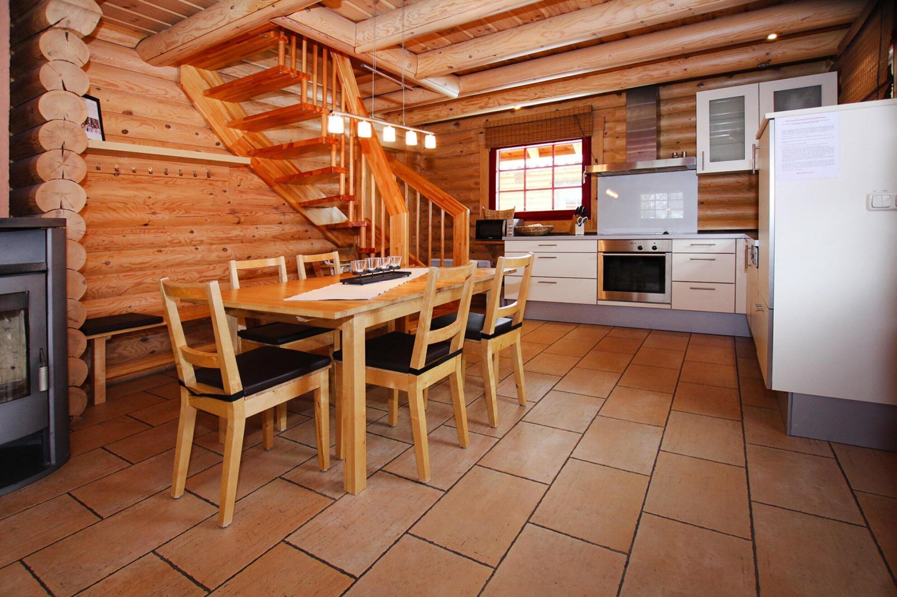 Interior de una cabaña de madera con mesa de comedor, sillas, una zona de cocina, chimenea y una escalera de madera.