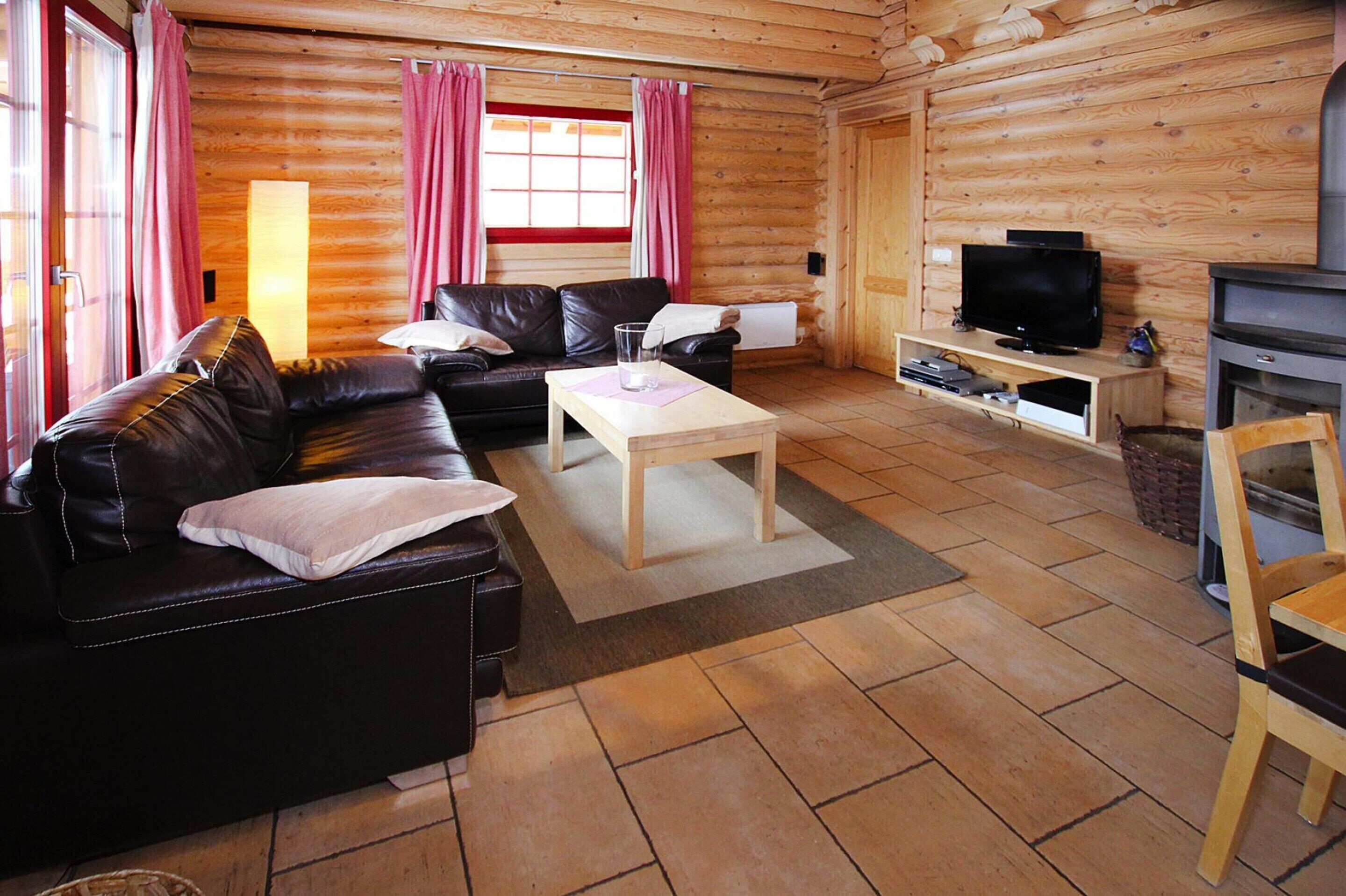 Interior de una cabaña de troncos con un sofá de cuero, una mesa de centro de madera, un televisor sobre un mueble bajo y una chimenea.