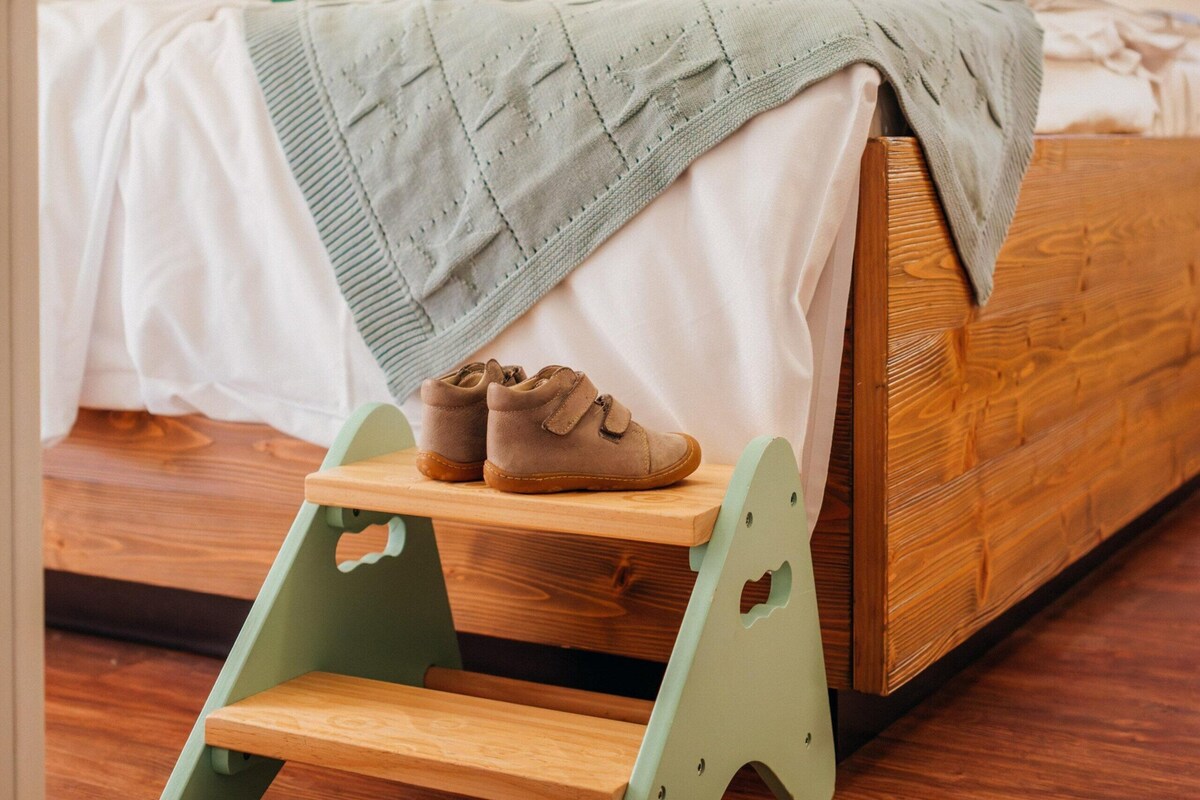 Un lit en bois pour enfant, avec un petit tabouret vert, un ours en peluche et des blocs de construction colorés posés sur le lit.
