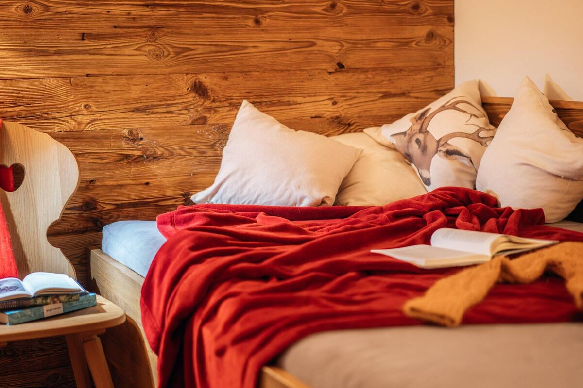 Cama con cabecera de madera, con una colcha y almohadas rojas, una mesita de noche con libros y una silla con un cojín con forma de corazón.