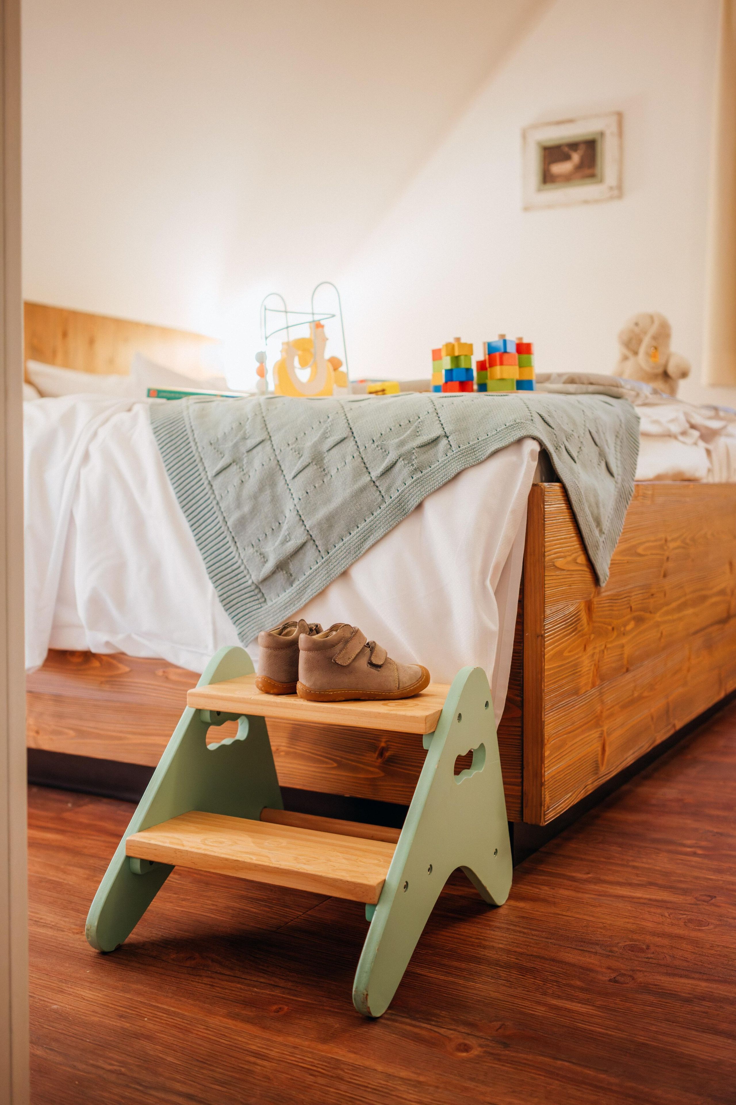 Un letto in legno con testata verde, un paio di scarpe marroni su un gradino di legno e mattoncini colorati sul letto.