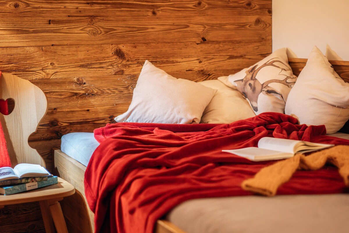 A wooden headboard bed with a red blanket and pillows, a small bedside table with books, and a chair with a heart-shaped cushion.