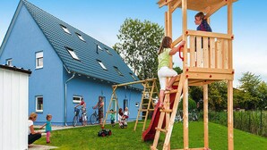 Un’area giochi con una struttura in legno per arrampicarsi e scivoli, sullo sfondo di una casa blu con tetto spiovente.