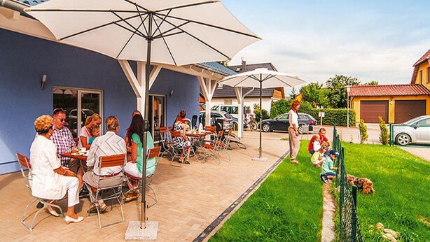 Un'area patio esterna con tavoli e sedie sotto grandi ombrelloni, circondata da un prato curato e una recinzione.