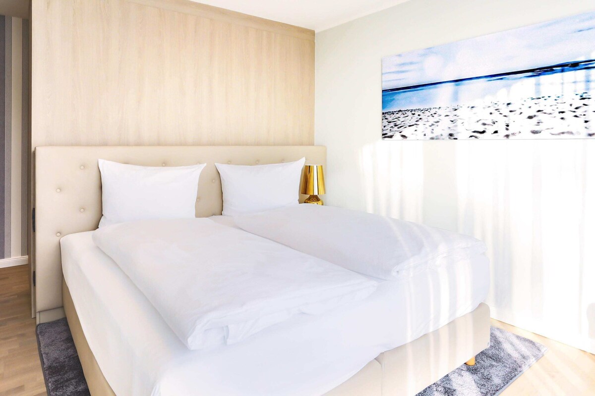 A modern hotel room with a large bed, a wooden headboard, and a painting of a beach scene.