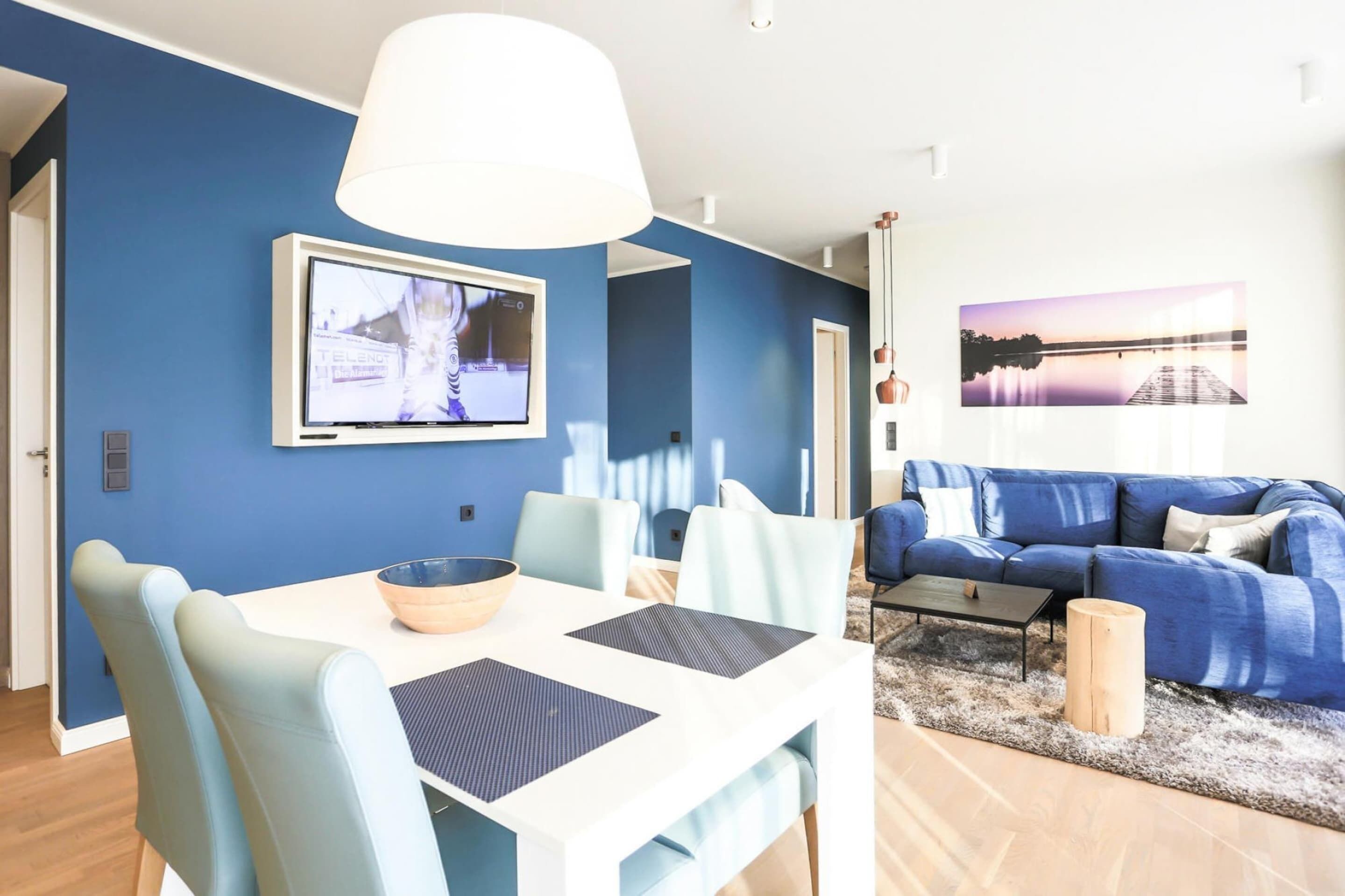 Un salon moderne avec un canapé bleu, une table à manger blanche avec des chaises bleues, un téléviseur fixé au mur et une grande fenêtre donnant sur un quai et un coucher de soleil.