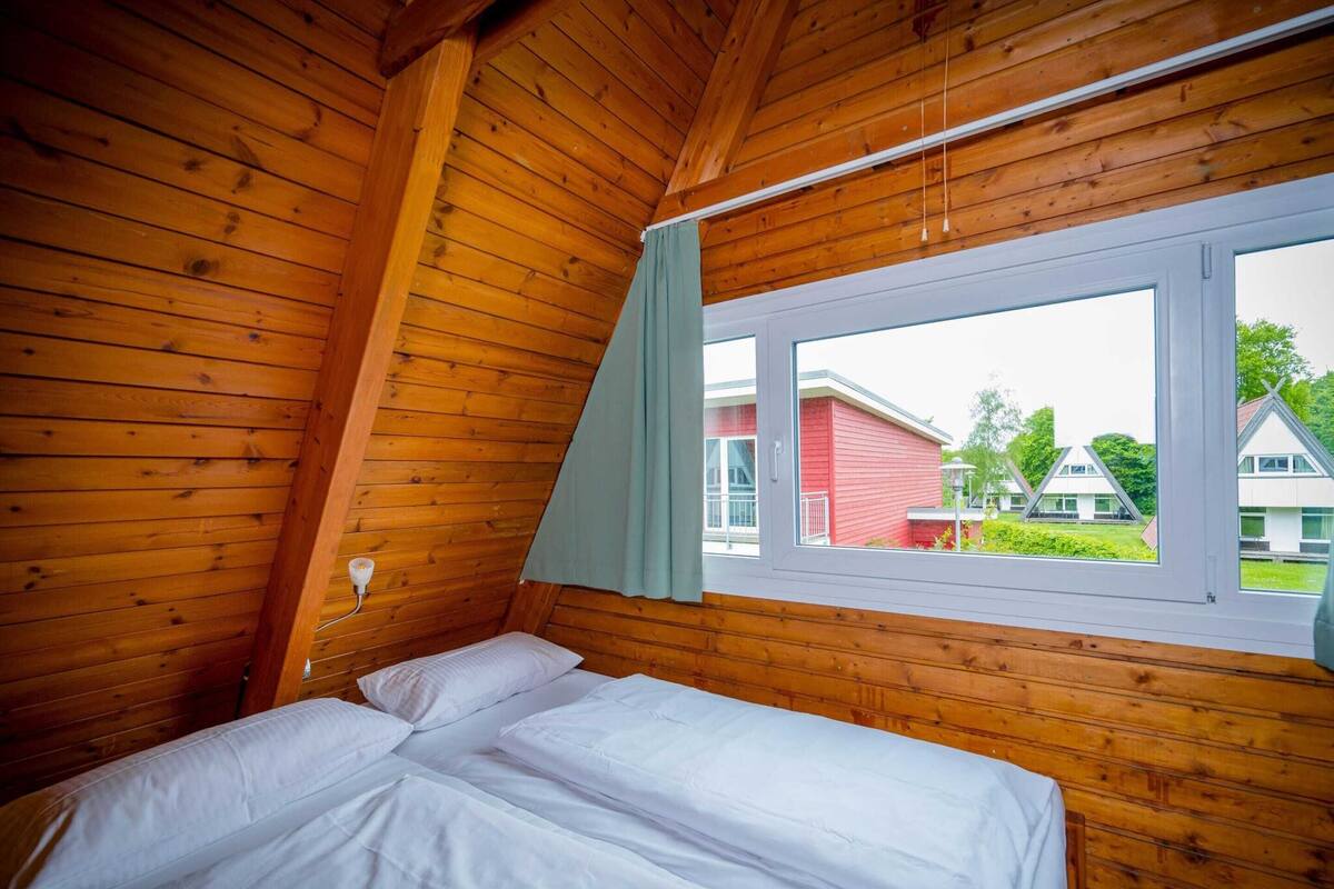 A wooden cabin room with a bed, a window, and a view of houses outside.