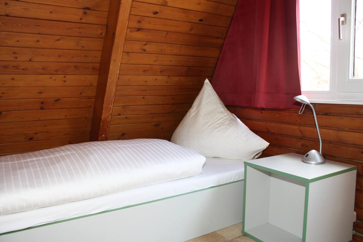 Une chambre dans un chalet en bois, avec un lit, une table de chevet et une fenêtre ornée d’un rideau rouge.