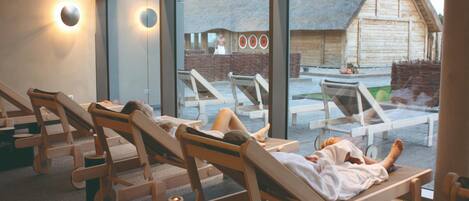 A spa area with wooden loungers, a glass wall, and a view of a rustic building.