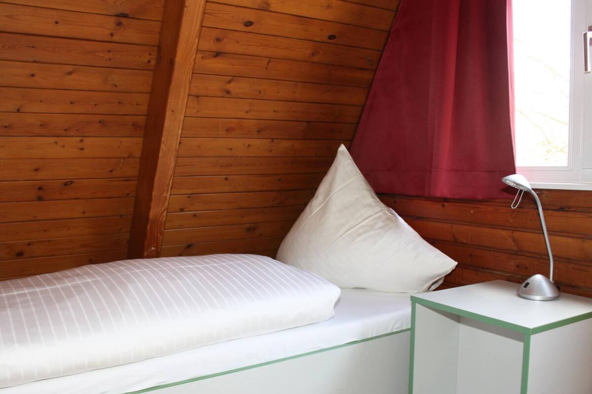 Une petite chambre avec un lit simple, des murs en bois, un lit blanc, une table de chevet à cadre vert et un rideau rouge.