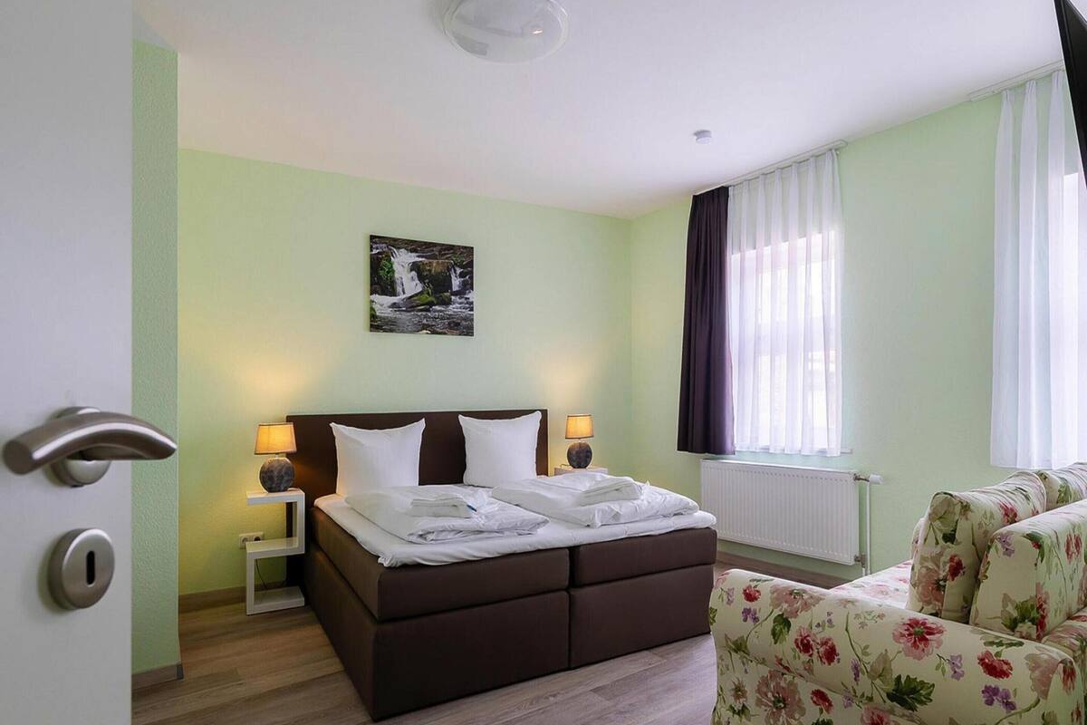 A hotel room with a bed, a chair, a nightstand, a lamp, a picture on the wall, and a window with curtains.