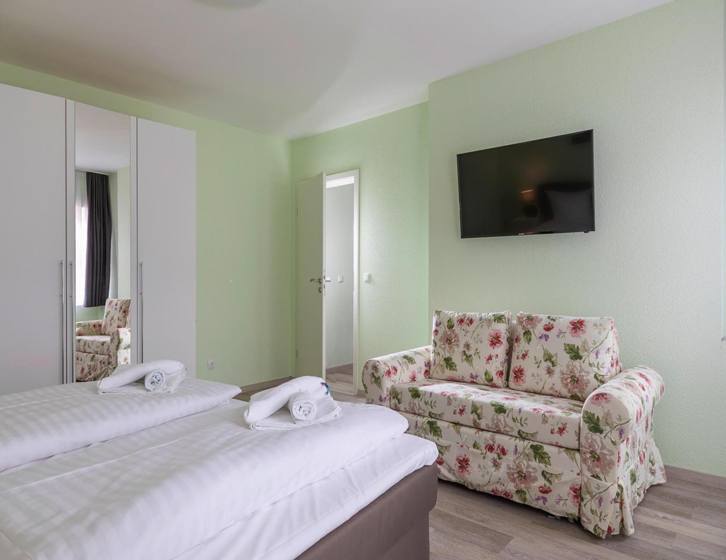 A hotel room with two beds, a floral sofa, a TV mounted on the wall, and a door leading to another room.